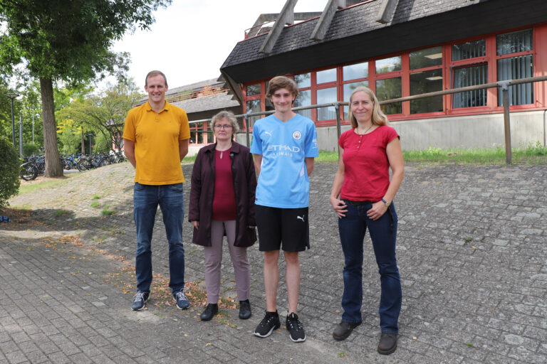 Christian Vennemann (Lehrer und Projektkoordinator), Claudia Leyers (Leiterin des Fachbereichs Umwelt und Klimaschutz der Stadt Osnabrück), Fynn Wekenborg (Schüler) und Maxi Hase (Projektkoordinatorin im Fachdienst Umweltplanung der Stadt) stehen vor einer Fläche, die bald entsiegelt und bepflanzt werden soll. / Foto: Simon Vonstein