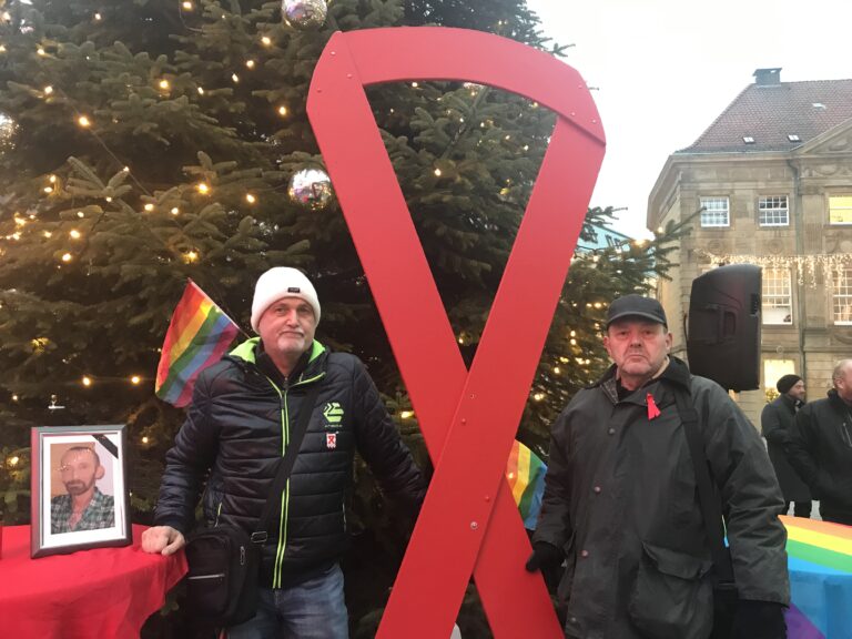 Martin Wellmann und Andreas Niemann mit großer Aidsschleife am Nikolaiort. Neben ihnen das Bild von ihrem Freund Alfred. / Foto: Burrichter