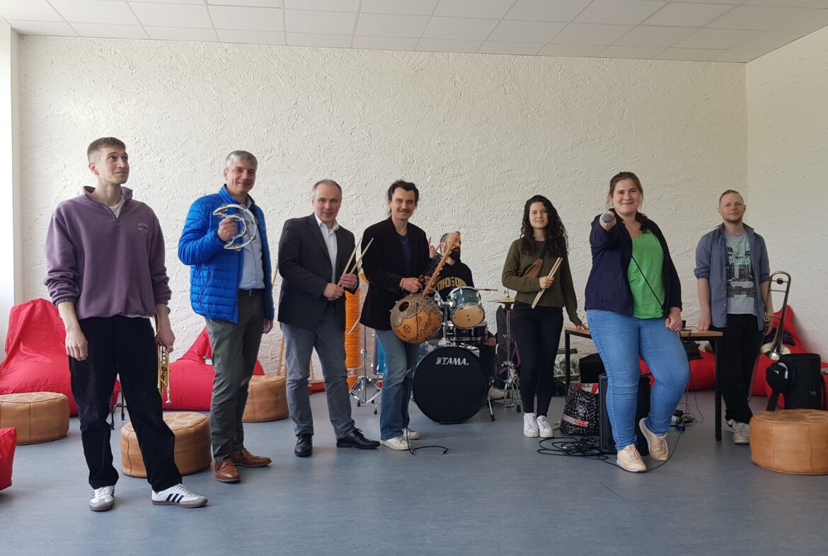 Die Musikerinnen und Musiker der "Banda Communale" mit Robert Klask (Fachgebietsleitung Aufnahme und Soziales und stellvertretende Standortleitung der LAB Sedanstraße, zweiter von links), Michael Prior (Vorstandssprecher der Friedel & Gisela Bohnenkamp-Stiftung, dritter von links) und Therese Sextro (Projektleiterin und Verantwortliche der Bildungsarbeit bei Exil e.V., zweite von rechts). / Foto: Rykov