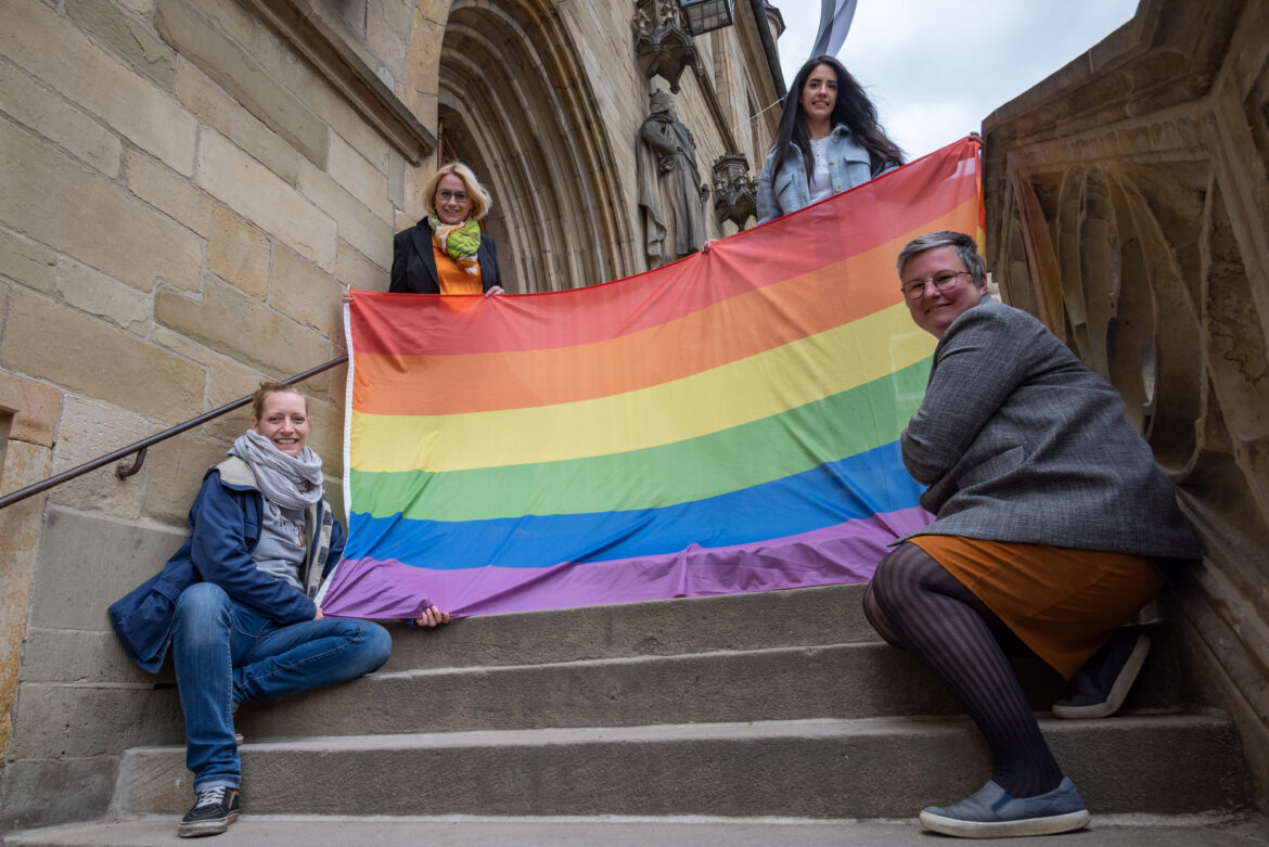 Oberbürgermeisterin Katharina Pötter (oben links), Heba Najdi vom Antidiskriminierungsbüro der Stadt Osnabrück (oben rechts), Ann Kristin Schneider, stellvertretende Gleichstellungsbeauftragte und Patricia Heller, Gleichstellungsbauftragte, mit der Regenbogenflagge, die am Dienstag, 17. Mai, an der Stadtbibliothek weht. / Foto: Monika Nestmann
