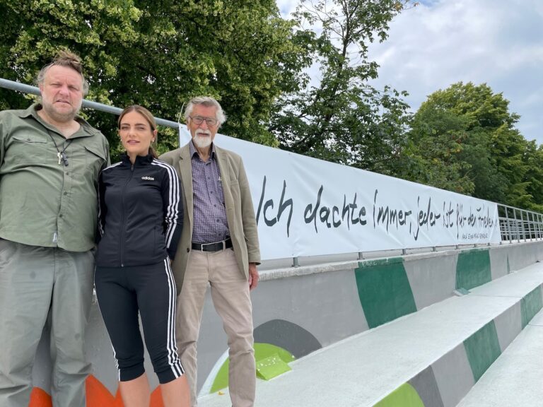 (von links) Künstler Volker-Johannes Trieb, Moskaubadleiterin Kerstin Klotz und Reinhold Mokrosch von der Remarque-Gesellschaft vor dem Banner auf der Tribüne des Moskaubads. / Foto: Stadtwerke Osnabrück