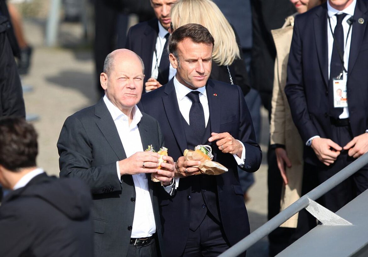 Bundeskanzler Olaf Scholz und Frankreichs Präsident Emmanuel Macron essen ein belegtes Brötchen