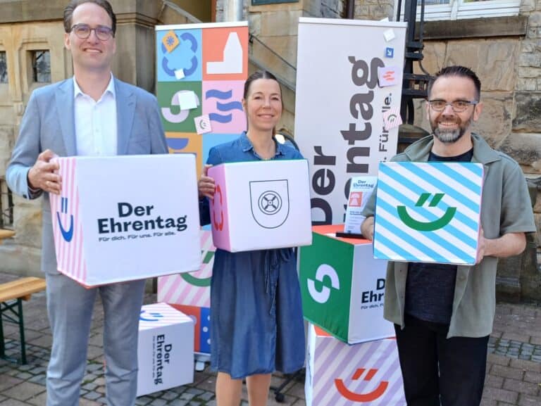 Am Rande der Eröffnung des 43. Meller Geranienmarktes warben (von links) Raphael Stock, Malina Kruse-Wiegand und Uwe Plaß für den Ehrentag, der am 23. Mai 2026 unter dem Leitgedanken „Für dich. Für uns. Für alle“ begangen wird./Foto: Stadt Melle