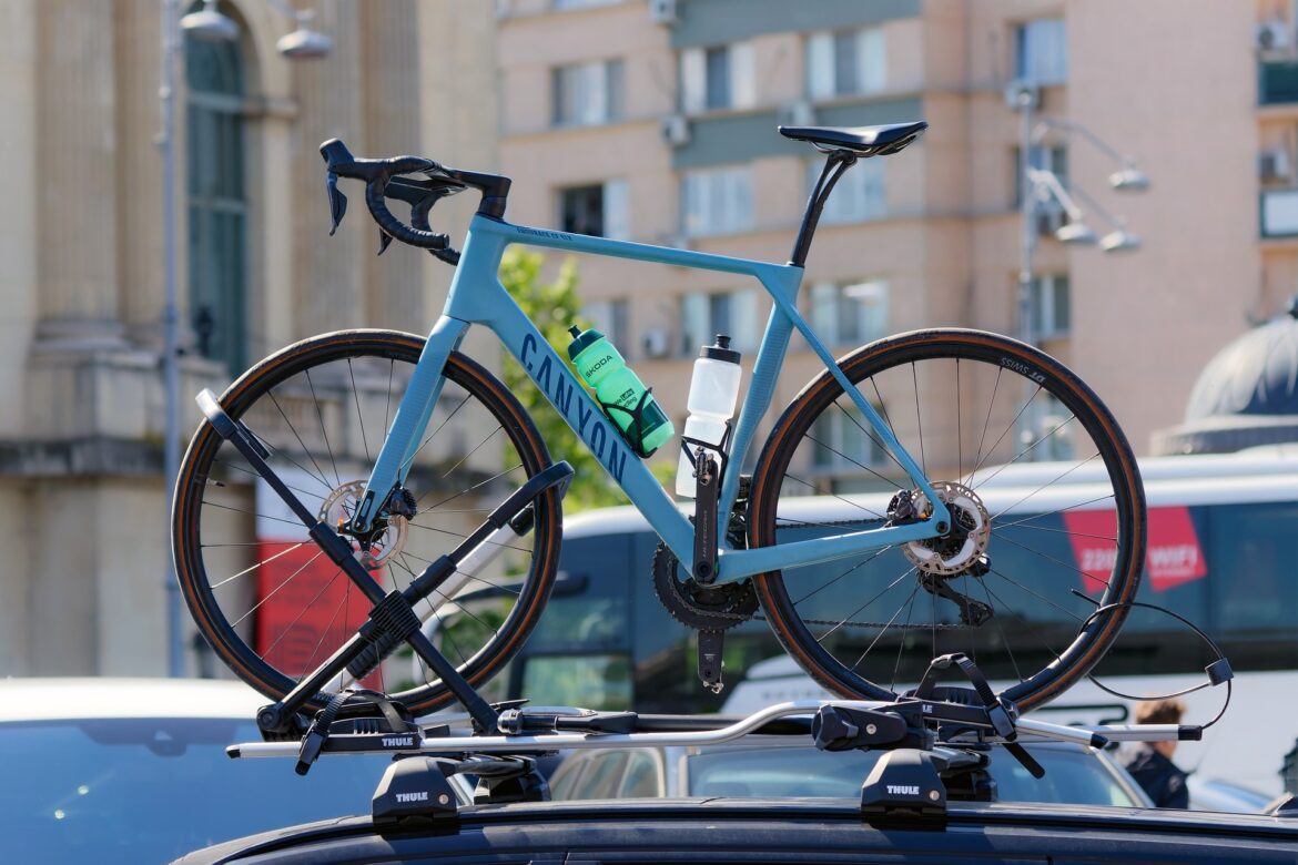 Ein hellblaues Rennrad mit zwei Trinkflaschen ist auf einem Fahrradträger auf dem Dach eines Autos montiert. Im Hintergrund sind unscharfe Stadtgebäude zu sehen.
