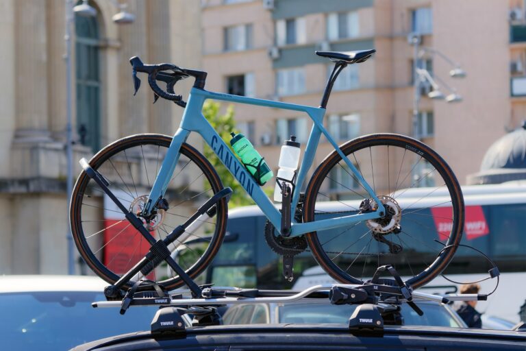Ein hellblaues Rennrad mit zwei Trinkflaschen ist auf einem Fahrradträger auf dem Dach eines Autos montiert. Im Hintergrund sind unscharfe Stadtgebäude zu sehen.