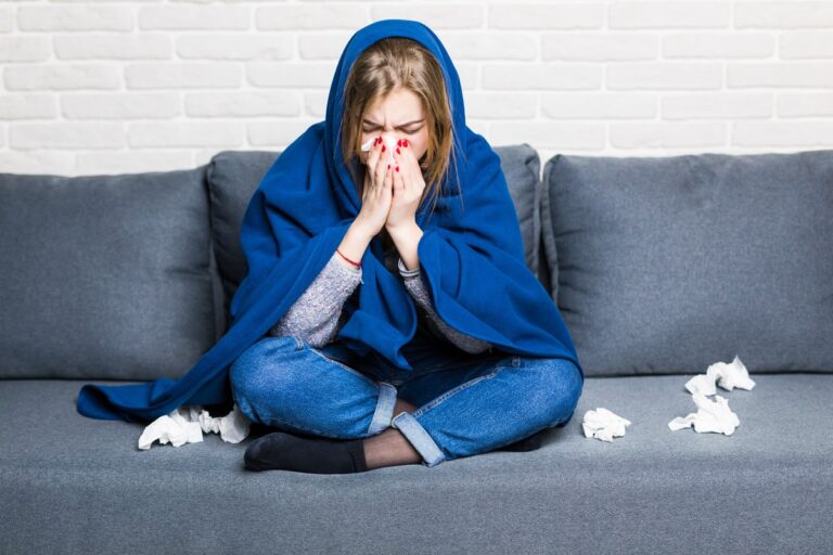 Erkältete junge Frau mit blauer Decke putzt sich auf dem Sofa die Nase.