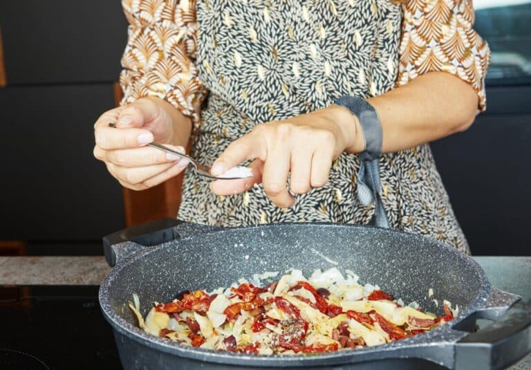 A person is adding spices with a spoon to a frying pan filled with sauted onions and sundried tomatoes
