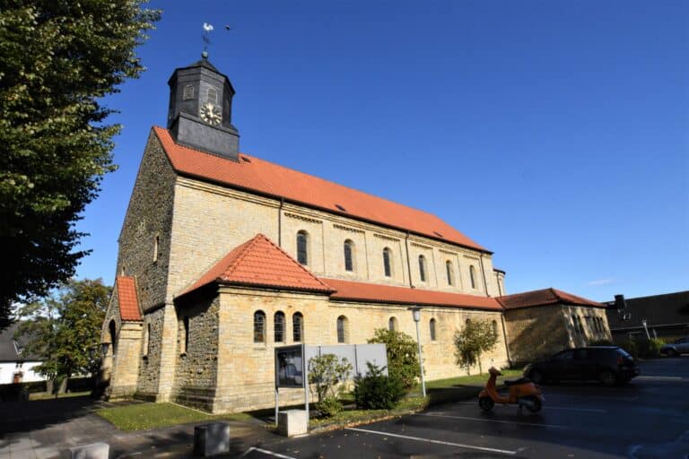 Backsteinrote Dächer und heller Natursteinbau der Liebfrauenkirche mit schwarzem Dachreiter und Uhr unter blauem Himmel, davor ein Parkplatz