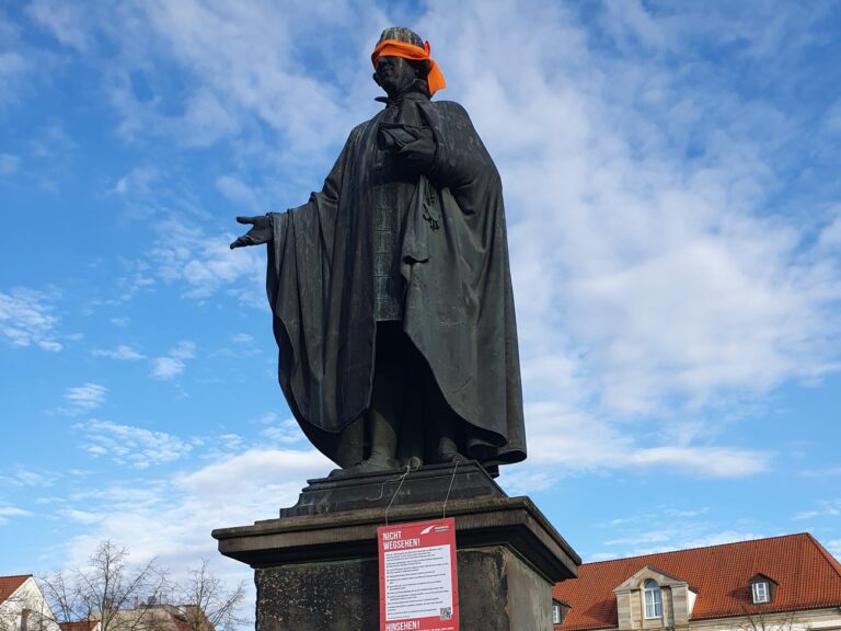 Justus-Möser-Denkmal in Osnabrück mit orangefarbener Augenbinde und Plakat der Seebrücke