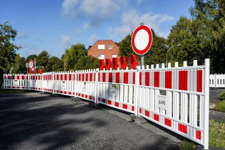 Weiß-roter Bauzaun mit Warnleuchten sperrt eine Straße, dahinter ein Verbotsschild für Fahrzeuge aller Art, ein Backsteinhaus und Bäume unter blauem Himmel