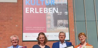 Vor dem acht Meter hohen Banner am Kreishaus werden Objekte rund um das Jubiläum des Bersenbrücker Museums präsentiert (von links): Kulturbüroleiter Burkhard Fromme mit einem Plakat für das Theater „Vorhang auf!“, Museumsleiterin Katharina Pfaff mit einem Portrait von Museumsgründer Rothert, Kreisrat Matthias Selle mit einem Aquarell Theodor Doebners und Museumspädagogin Claudia Stuckenberg mit Museumsführern aus drei Epochen. / Foto: Henning Müller-Detert